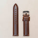 Brown leather watch strap with silver buckle on a beige background
